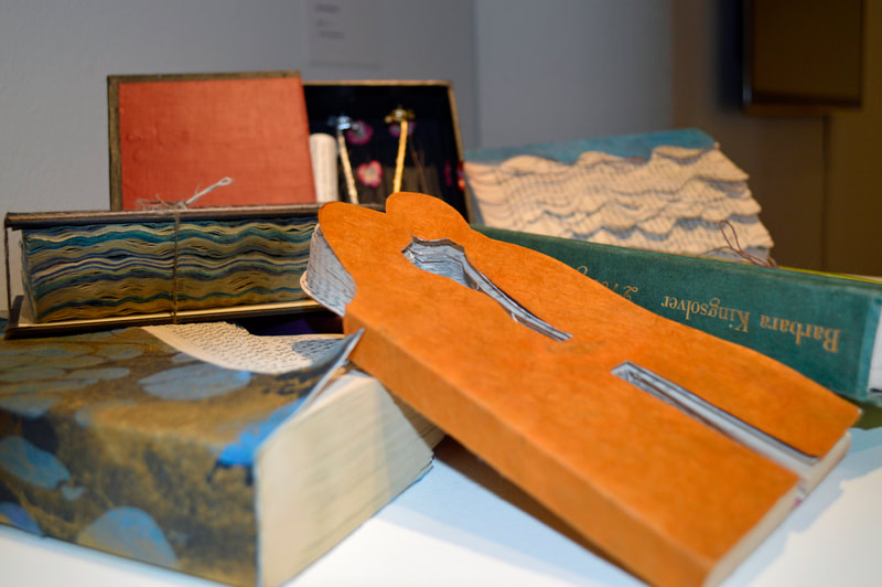A disorganized pile of artist's books and boxes on top of a podium. Most of the books are covered with decorative paper. One is cut into the shape of a couple kissing. An open box reveals two giraffe statuettes, flowers, and a paper scroll.