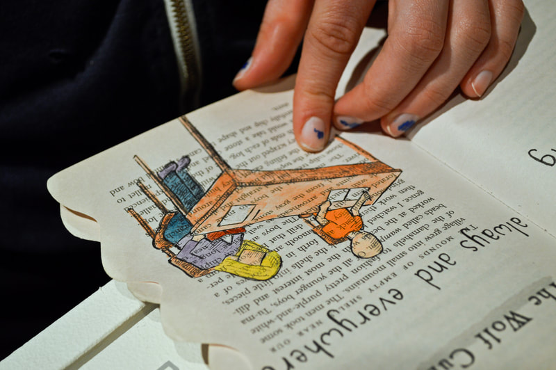 A hand with chipped nail polish on its fingernails holds a book open. An ink and watercolor drawing of two children sitting at a desk is layered over the book's text. 