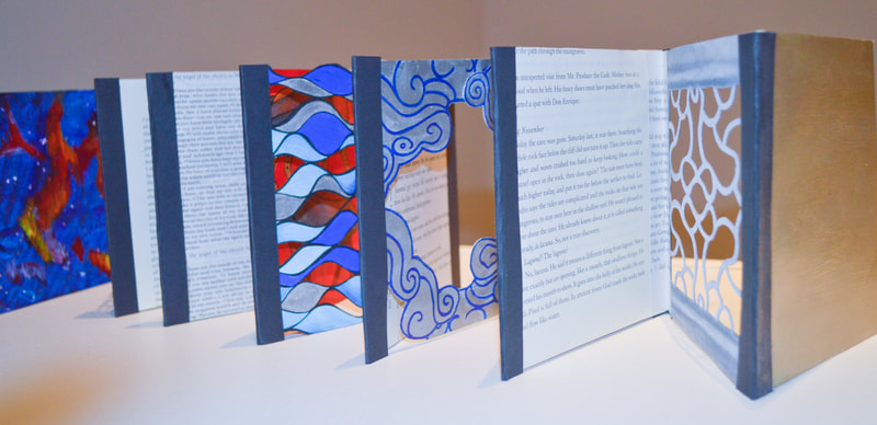 Portion of an accordion book. The panels in view include silver and blue abstract waves with sections cut out; swirly, abstract clouds around a central cut-out; a cut-paper version of a Chinese garden window, and two pages from books.