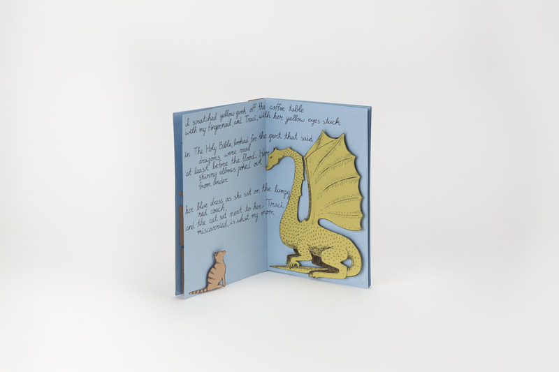 A pop-up book propped open. An illustration of a cat faces an illustration of a dragon. A piece of a poem is handwritten between the two illustrations.