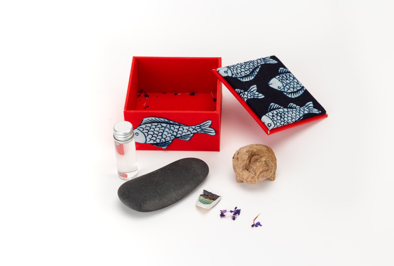 A red box decorated with fish stands with its lid open. A small bottle, a hand-sized rock, a wood carving in the shape of a fox, a ceramic chip, and small purple flowers are arrayed in front of it.