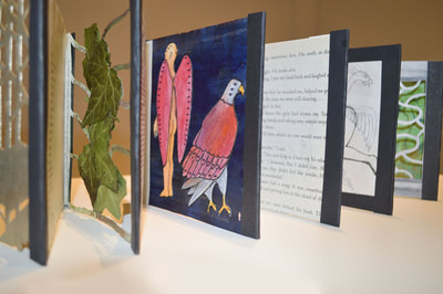Portion of an accordion book. The panels in view include a paper lattice with pressed ivy leaves stacked on top of it, an illustration of a man with wings and an eagle standing back to back, and a page from a book.