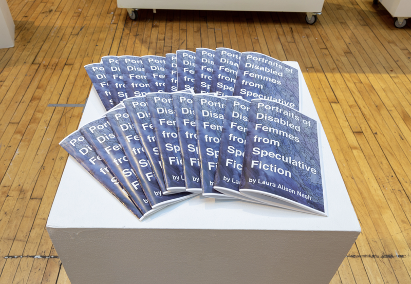 Several copies of a booklet spread on top of a white pedestal, which stands on a wooden floor. The booklet is titled "Portraits of Disabled Femmes from Speculative Fiction by Laura Alison Nash," in white lettering on a purplish cover.